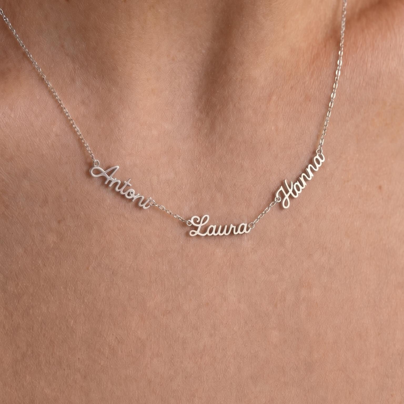 A close-up of a delicate silver necklace worn on a person's neck, featuring personalized script names 'Antoni', 'Laura', and 'Hanna' spaced along a fine chain.