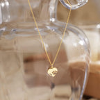 A gold chain necklace with a small heart-shaped pendant engraved with a paw print, hanging in front of a blurred clear glass vase.