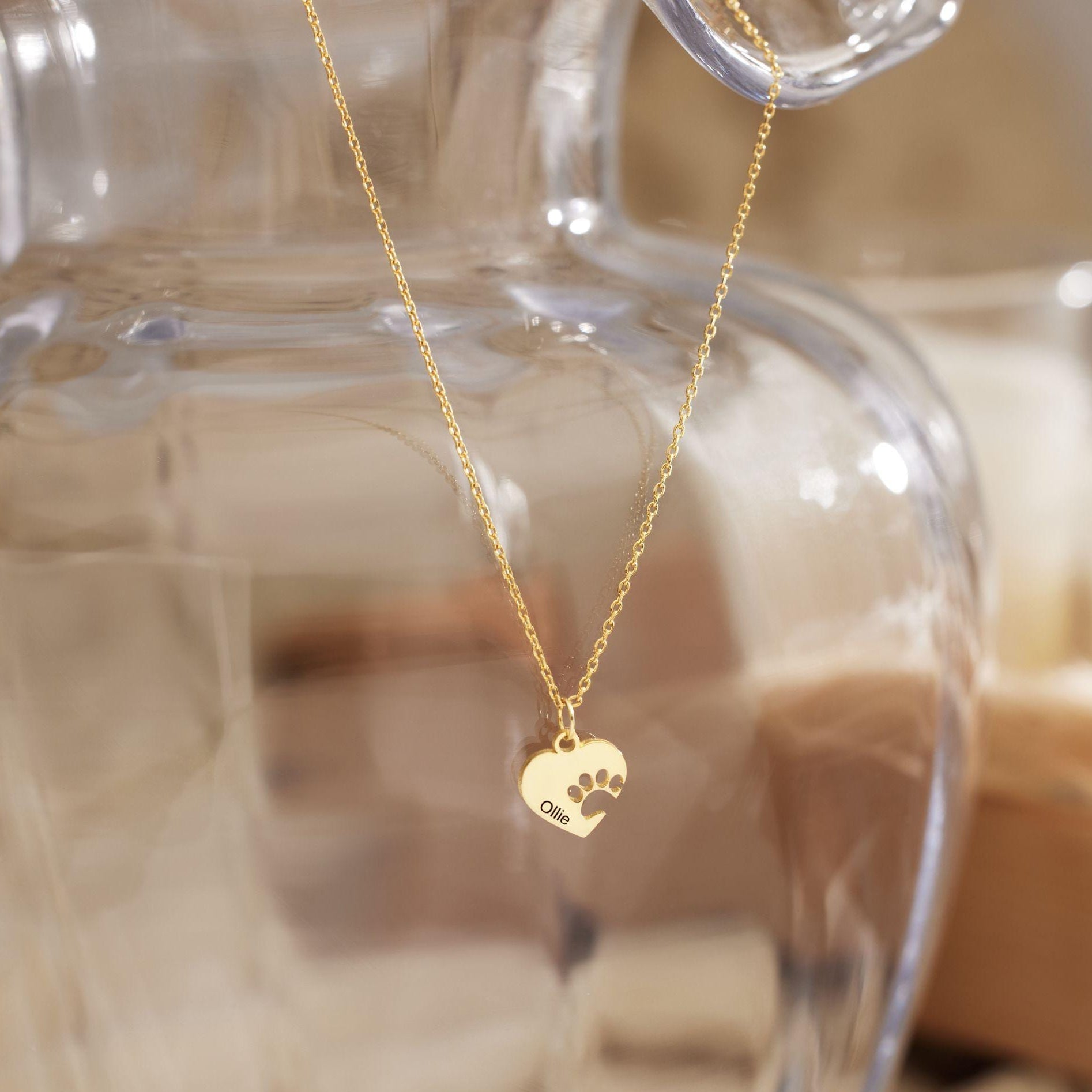 A gold chain necklace with a small heart-shaped pendant engraved with a paw print, hanging in front of a blurred clear glass vase.