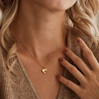 A close-up lifestyle shot of a model wearing a delicate gold chain necklace with a small, polished heart-shaped pendant. The model has blonde hair and is wearing a brown knit cardigan, with her hand resting near her chest.