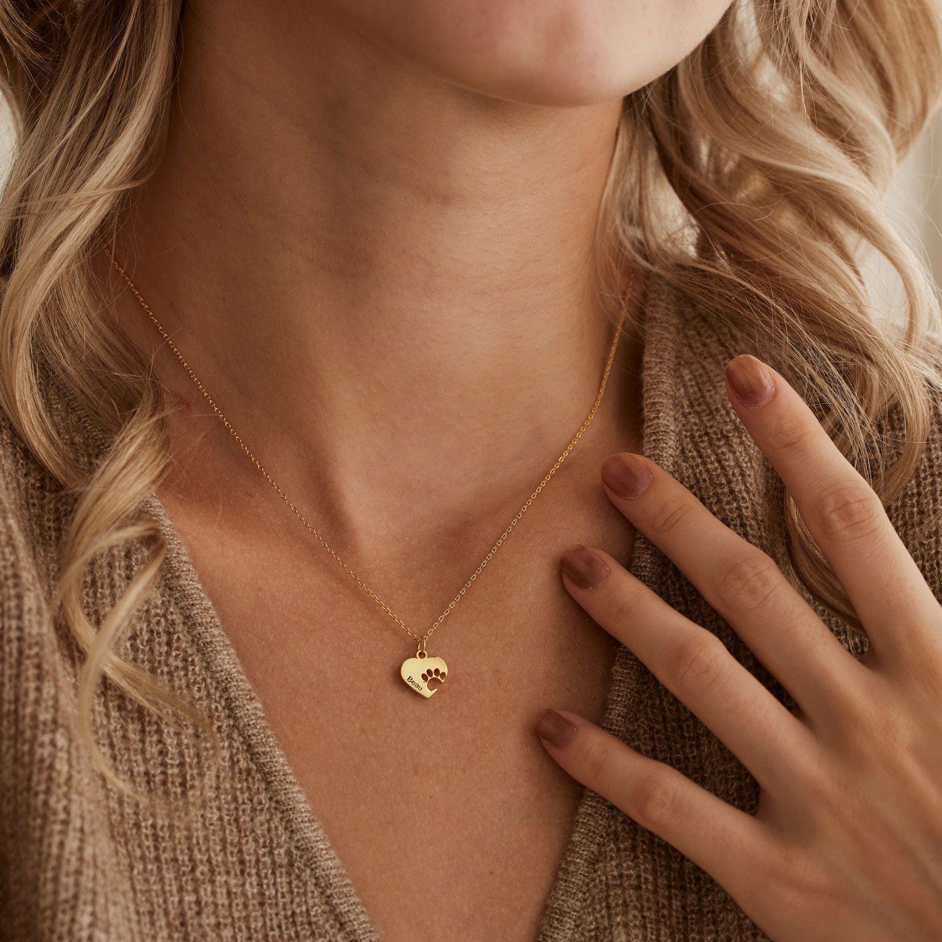 A close-up lifestyle shot of a model wearing a delicate gold chain necklace with a small, polished heart-shaped pendant. The model has blonde hair and is wearing a brown knit cardigan, with her hand resting near her chest.