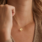 A close-up lifestyle shot of a model wearing a delicate gold chain necklace with a small heart-shaped pendant. The pendant features a cutout design of a small dog.
