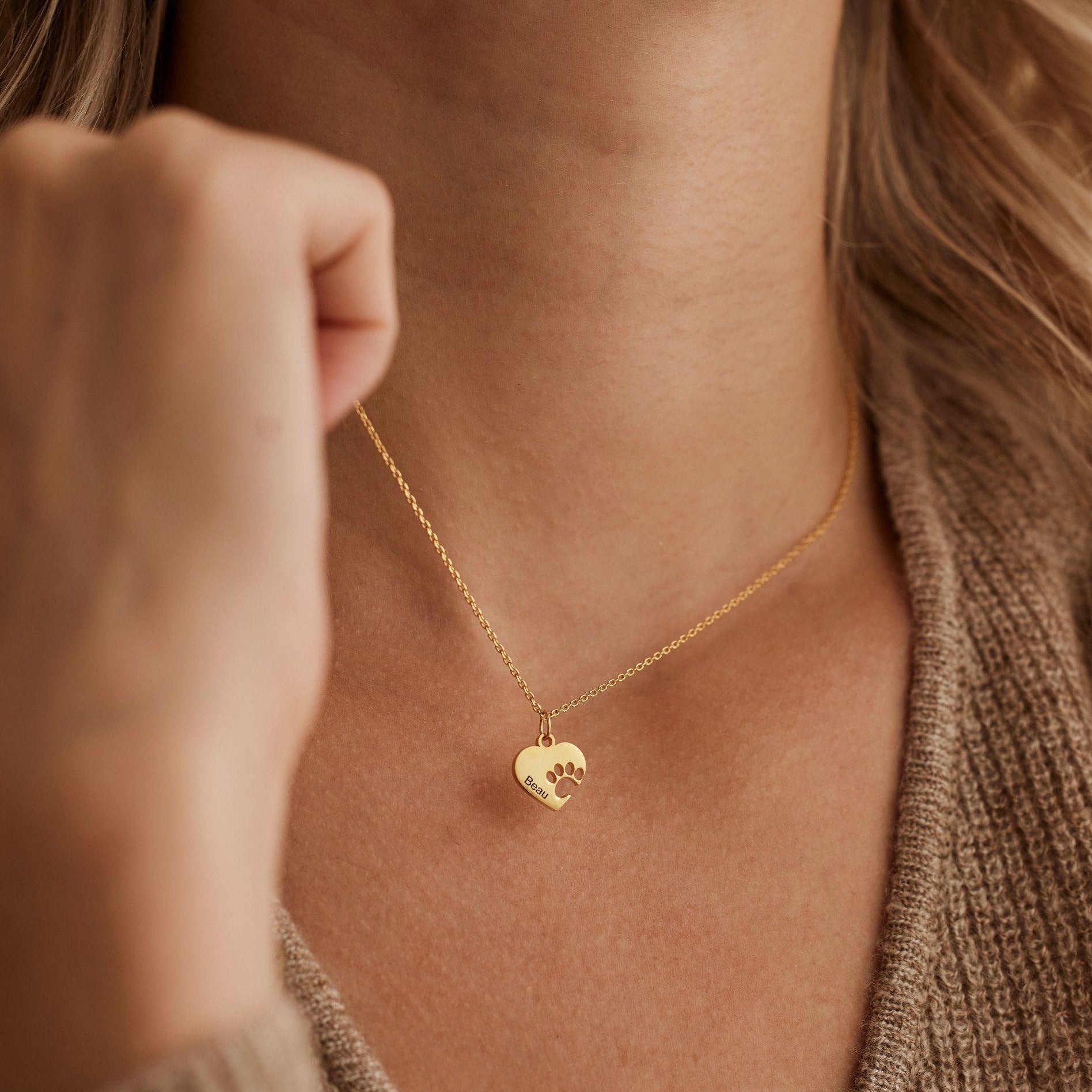 A close-up lifestyle shot of a model wearing a delicate gold chain necklace with a small heart-shaped pendant. The pendant features a cutout design of a small dog.