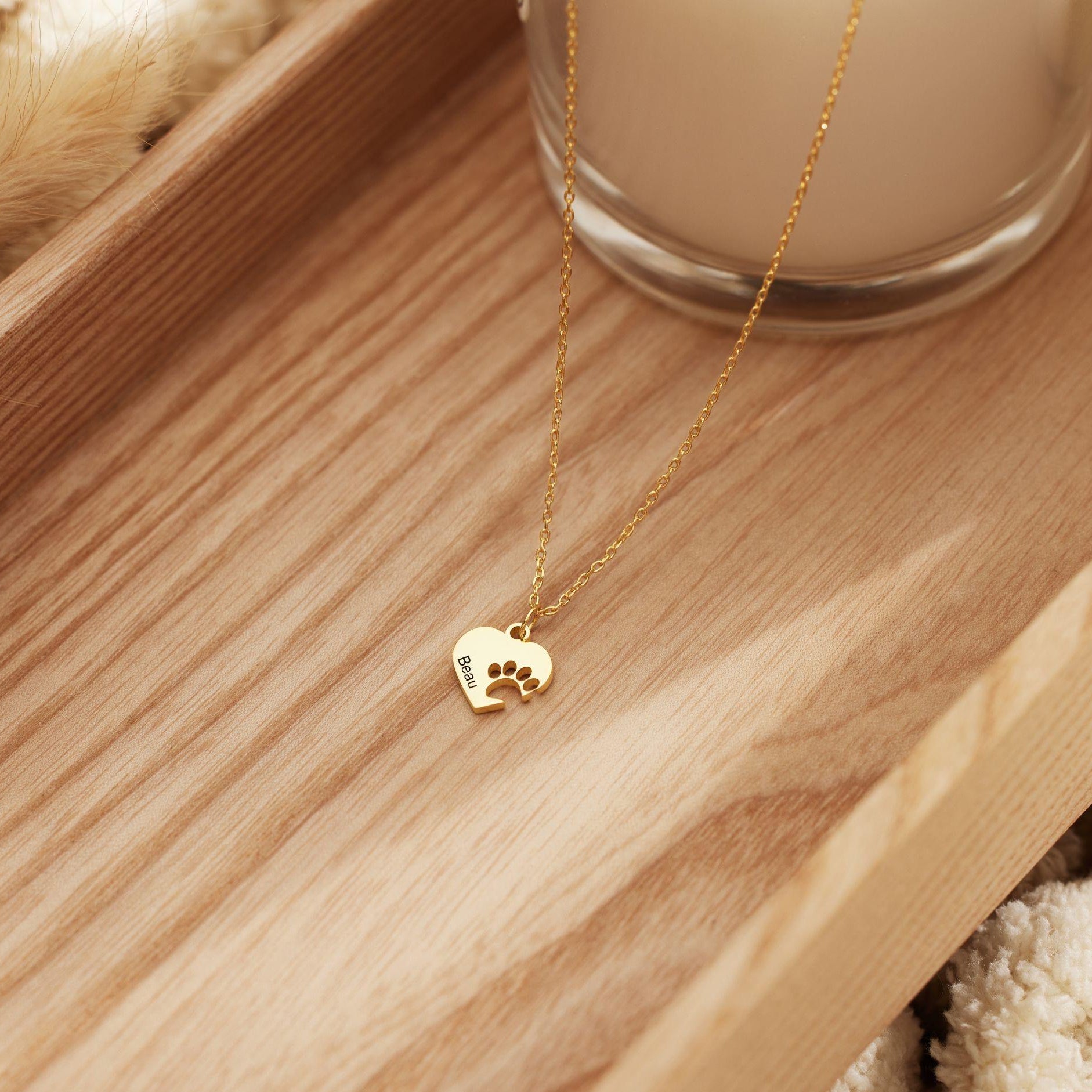A gold chain necklace with a small heart-shaped pendant featuring a paw print and the name 'Mochi', displayed on a wooden tray next to a glass candle.