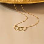 Gold family name necklace with three name hearts and a fine cable chain laid out on a beige surface.