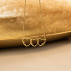 Personalised gold heart necklace with three engraved names, displayed on a gold tray against a beige backdrop.
