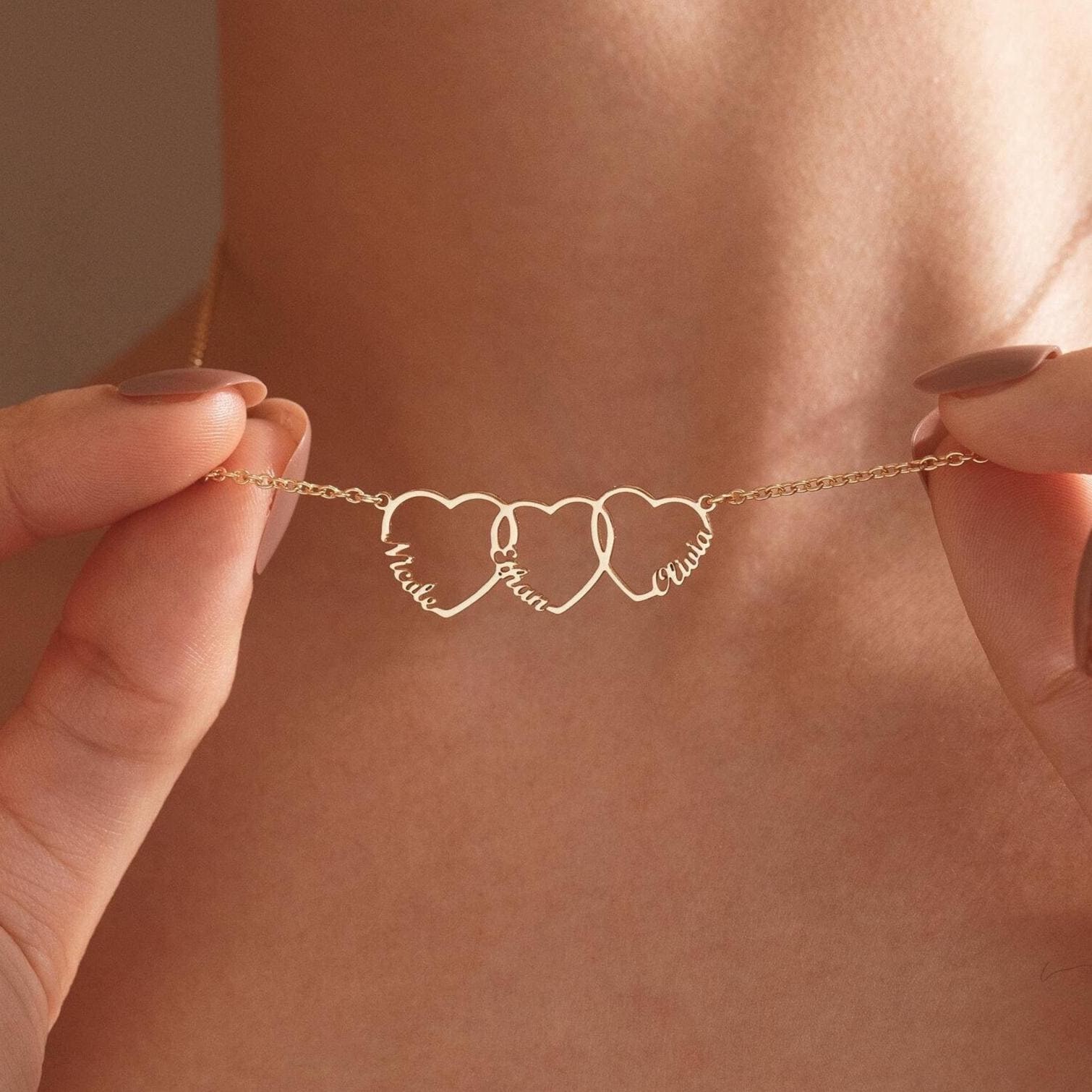Close-up of a gold family name necklace with three interlocking open hearts, each shaped around a child's name, held up against a woman's neck.