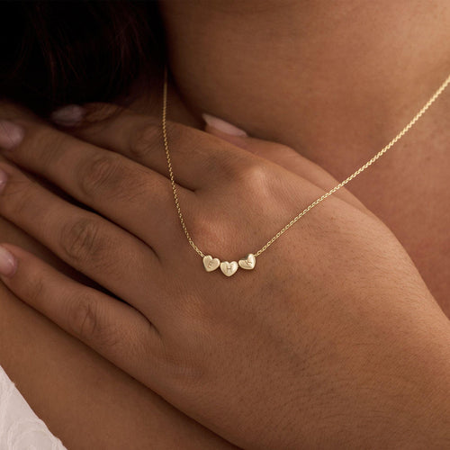 A close-up lifestyle shot of a delicate gold chain necklace with three small, individually-engraved heart charms, worn on a model's neck.